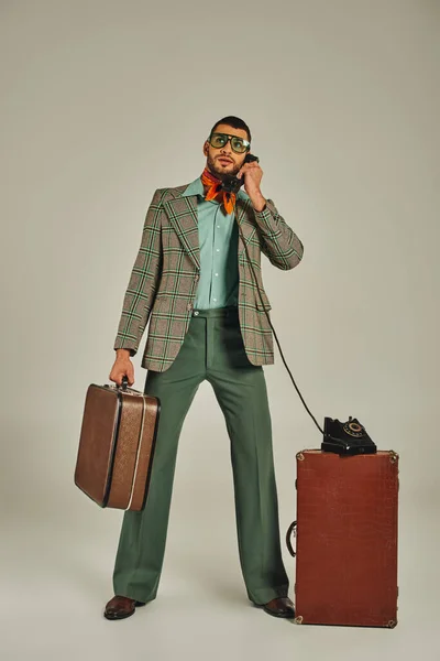 Uomo elegante in abbigliamento vecchio stile che tiene valigia vintage e parla su smartphone su grigio — Foto stock