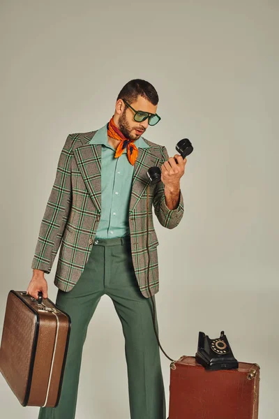 Uomo di tendenza in occhiali da sole che tengono valigia vintage e portatile sul telefono cordato su grigio, moda retrò — Foto stock