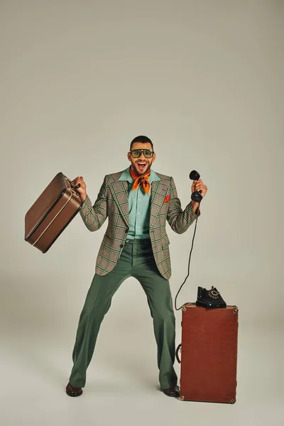 Homme ravi dans des vêtements de style rétro avec valise vintage et téléphone à cordon sur gris, pleine longueur — Photo de stock