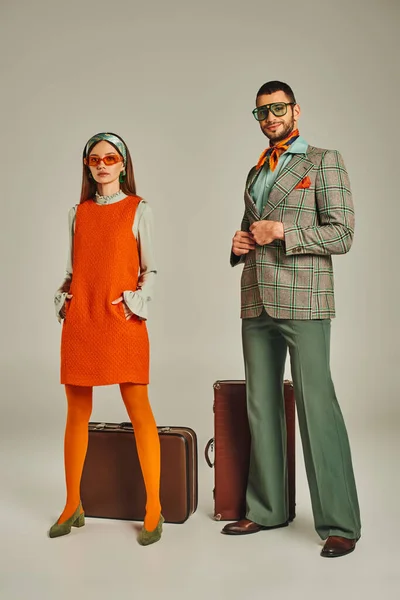 Donna in abito arancione e occhiali da sole vicino felice uomo in giacca a quadri e valigie vintage su grigio — Foto stock