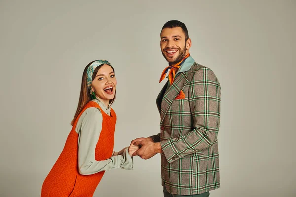 Young joyful couple in stylish vintage attire holding hands and looking at camera on grey backdrop — Stock Photo