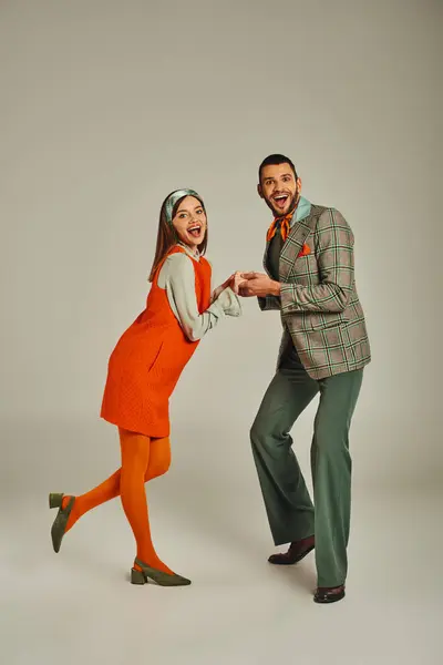 Full length of overjoyed couple in elegant retro attire looking at camera and holding hands on grey — Stock Photo