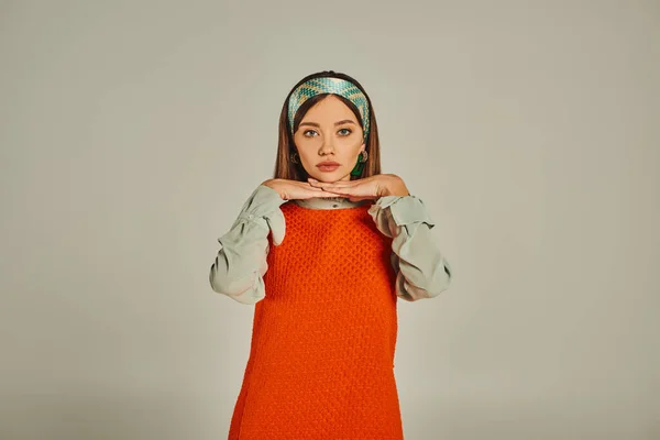 Giovane donna in fascia colorata e abito arancione posa con le mani sotto il mento su grigio, stile retrò — Foto stock