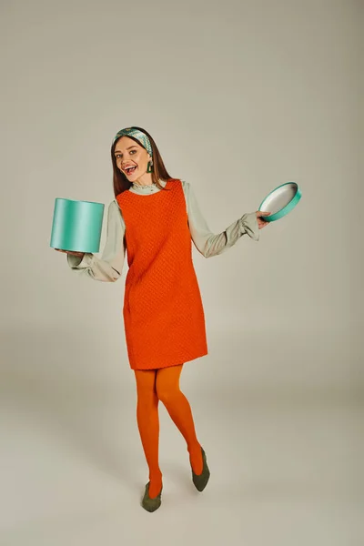 Volle Länge der Frau in orangefarbenem Retro-Stil Kleid mit Geschenkbox und Blick in die Kamera auf grau — Stockfoto