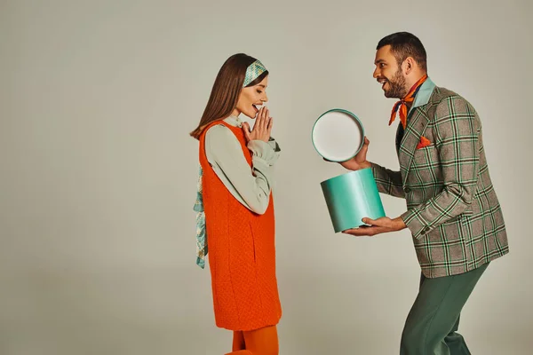 Moda uomo apertura confezione regalo vicino donna sorpresa in abito arancione su grigio, coppia di ispirazione retrò — Foto stock
