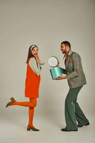 Stylish man holding gift box near overjoyed woman in orange dress on grey, retro-inspired fashion — Stock Photo