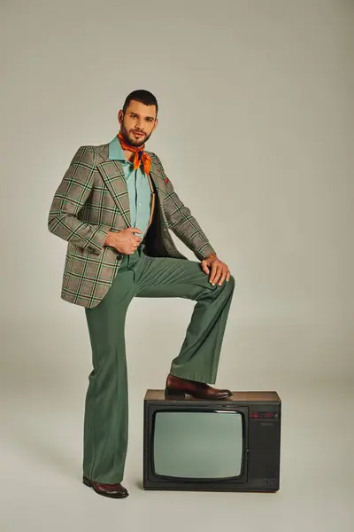 Confident man in stylish retro clothes stepping on vintage tv set and looking at camera on grey — Stock Photo