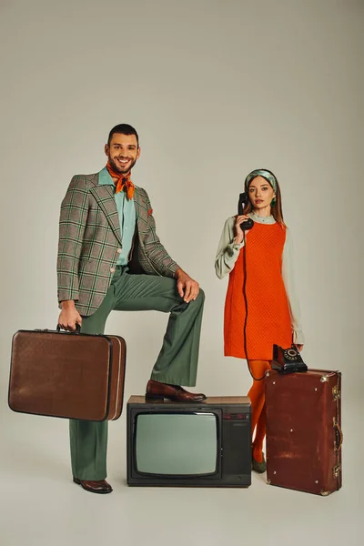 Femme de style rétro parlant sur le téléphone vintage près du téléviseur classique et homme heureux avec valise sur gris — Photo de stock