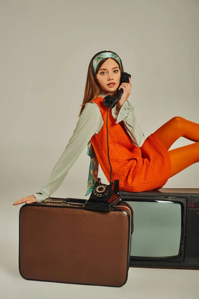 Очаровательная ретро-женщина сидит на винтажном чемодане и телевизоре, разговаривая по телефону на сером — стоковое фото