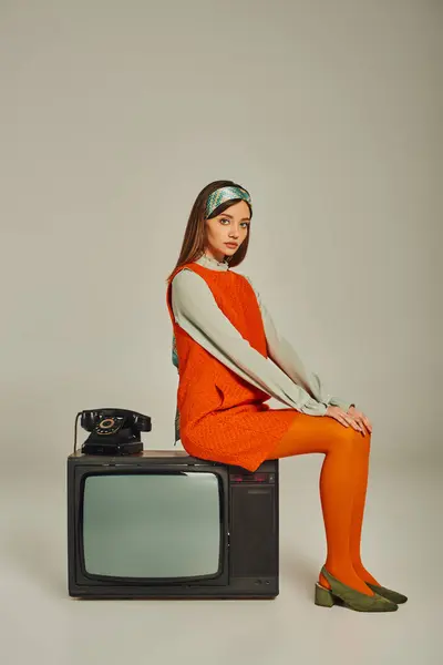 Модная женщина в ретро-стиле одежда сидит на винтажном телевизоре рядом с проволочным телефоном на сером, полная длина — стоковое фото