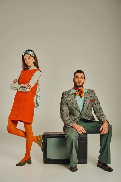 Женщина в оранжевом платье со сложенными руками рядом мужчина сидит на винтажном телевизоре на сером, ретро моде — стоковое фото