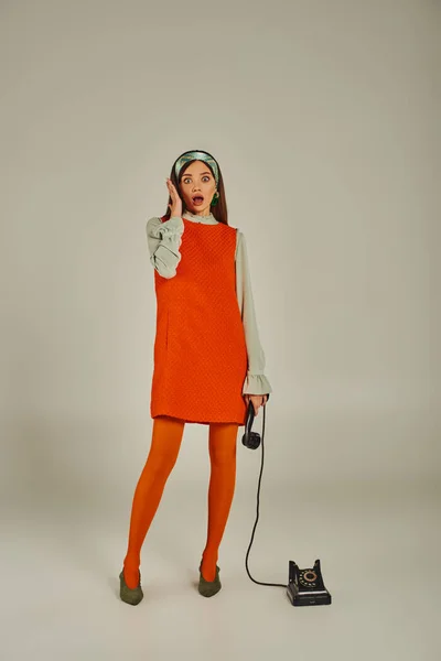 Шокированная женщина в стильном оранжевом платье держит телефон винтажного цвета на сером, полной длины — стоковое фото