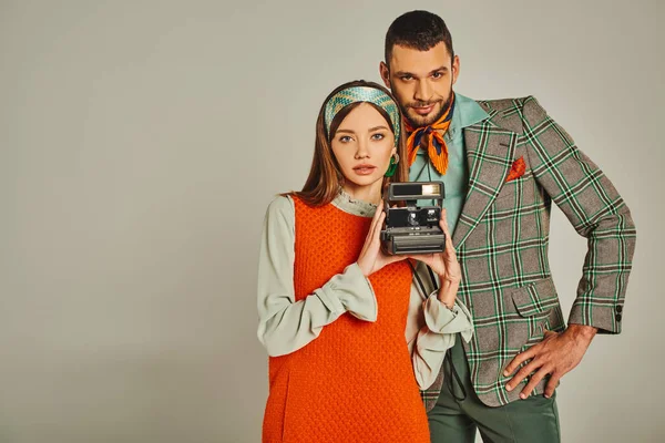 Jovem casal em traje elegante com câmera vintage olhando para a câmera em cinza, retro-inspirado moda — Fotografia de Stock