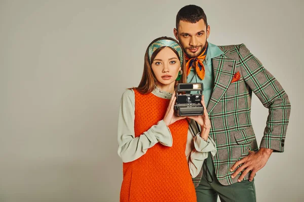 Pareja joven en traje elegante con cámara vintage mirando a la cámara en gris, moda de inspiración retro - foto de stock