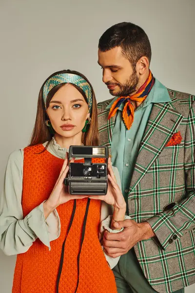 Homem elegante em jaqueta xadrez olhando para a mulher em vestido laranja segurando câmera vintage em cinza — Fotografia de Stock