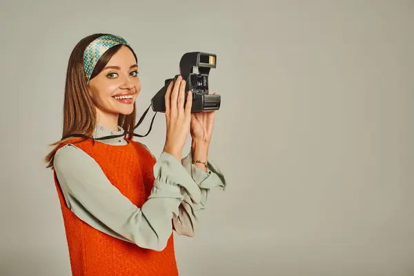 Mujer alegre en vestido naranja y diadema brillante tomando foto en la cámara vintage en gris, estilo retro - foto de stock