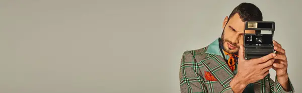 Модный мужчина в клетчатом пиджаке и ярком платке, фотографирующий на винтажную камеру на сером, баннере — стоковое фото
