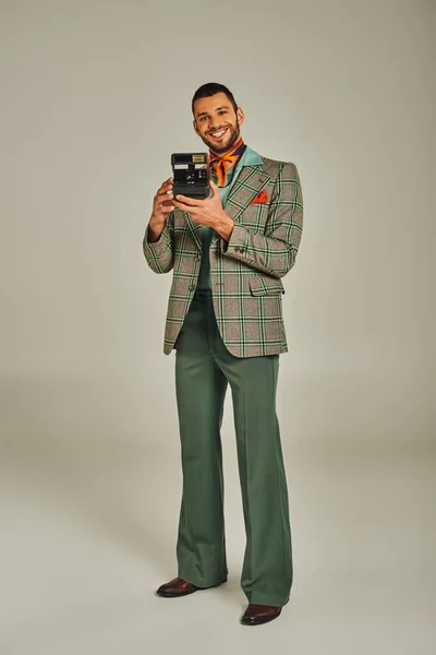 Longitud completa de hombre alegre en elegante atuendo retro de pie con cámara vintage sobre fondo gris - foto de stock