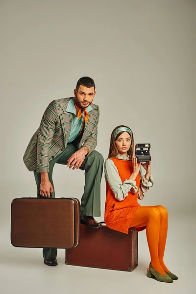 Woman in orange dress sitting on suitcase with vintage camera near retro style man on grey — Stock Photo
