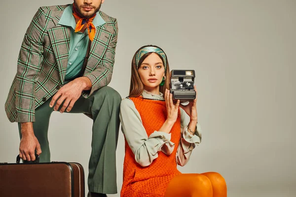 Woman in orange dress holding vintage camera near man with suitcase on grey, retro fashion — Stock Photo