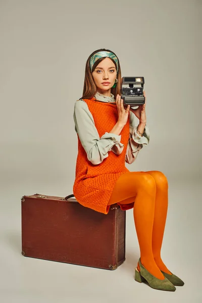 Femme à la mode assise sur une valise et prenant des photos sur un appareil photo vintage sur gris, voyageur à l'ancienne — Photo de stock