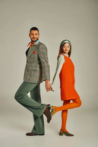 Feliz pareja en elegante traje vintage de pie espalda con espalda y tomados de la mano en gris, longitud completa - foto de stock