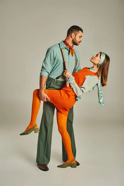 Vista lateral del hombre con estilo en tirantes coqueteando con la mujer en ropa brillante en gris, moda retro - foto de stock