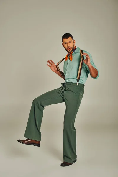Animado hip man em roupas vintage elegantes puxando suspensórios e olhando para a câmera no cinza — Fotografia de Stock