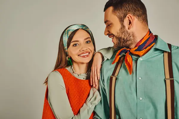 Joyful woman in orange dress looking at camera near man in suspenders on grey, retro-inspired style — Stock Photo