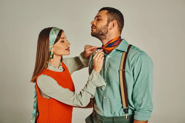Charming woman tying bright neckerchief on stylish man in suspenders on grey, vintage fashion — Stock Photo