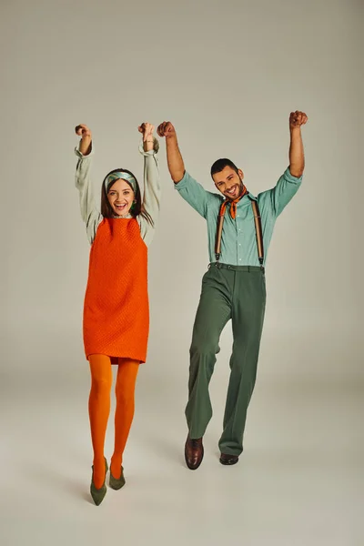 Couple gai en vêtements vintage dansant avec les mains levées sur gris, bonheur et vibes rétro — Photo de stock