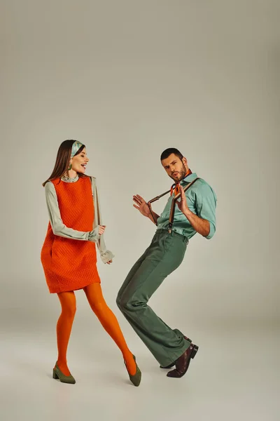 Hombre de moda tirando tirantes mientras baila cerca de mujer alegre en vestido naranja en gris, estilo retro - foto de stock