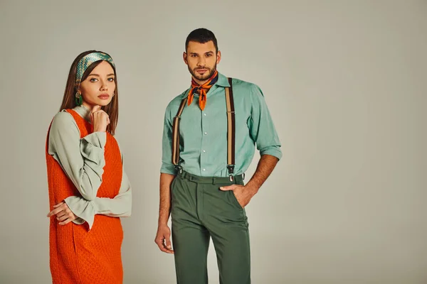 Confident man with hand in pocket near thoughtful woman in orange dress on grey, retro fashion — Stock Photo