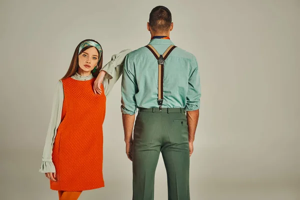 Young woman in orange dress looking at camera near trendy man in suspenders on grey, vintage style — Stock Photo