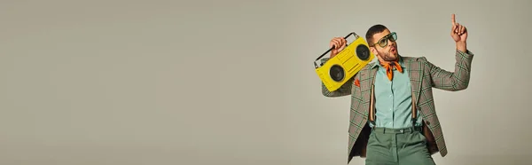 Excited stylish man in sunglasses holding vintage boombox, pointing up and dancing on grey, banner — Stock Photo