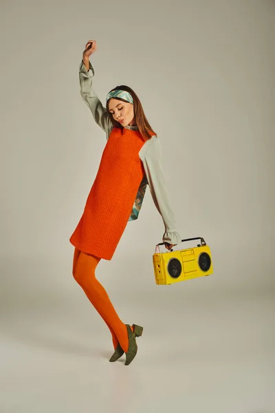 Mulher animada em vestido laranja dançando com boombox amarelo e levantou a mão em cinza, vibrações vintage — Fotografia de Stock