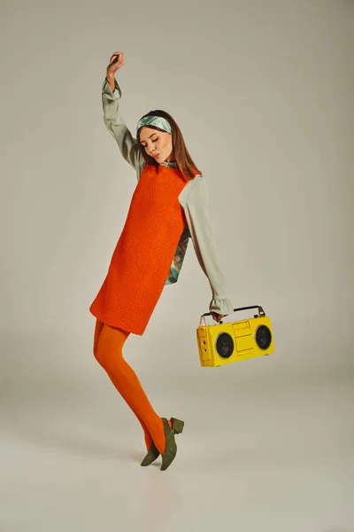 Excited woman in orange dress dancing with yellow boombox and raised hand on grey, vintage vibes — Stock Photo