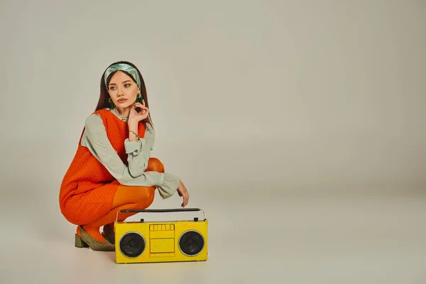 Femme réfléchie en robe orange écouter de la musique sur boombox jaune sur gris, style de vie rétro-inspiré — Photo de stock