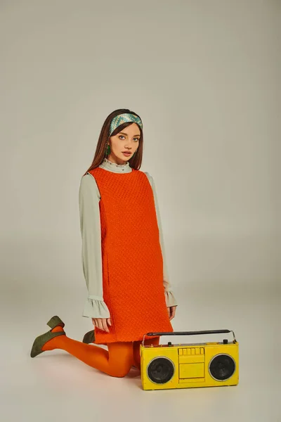 Jeune femme en robe orange vif et bandeau agenouillé près de boombox jaune sur gris, mode rétro — Photo de stock