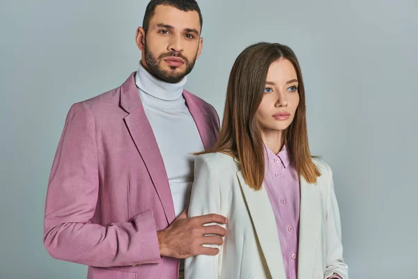 Uomo sicuro di sé in blazer lilla guardando la fotocamera vicino alla donna in abito bianco su grigio, moda business — Foto stock