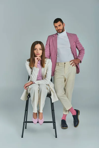 Homem confiante em blazer lilás com a mão no quadril perto da mulher em terno branco sentado na cadeira no cinza — Fotografia de Stock