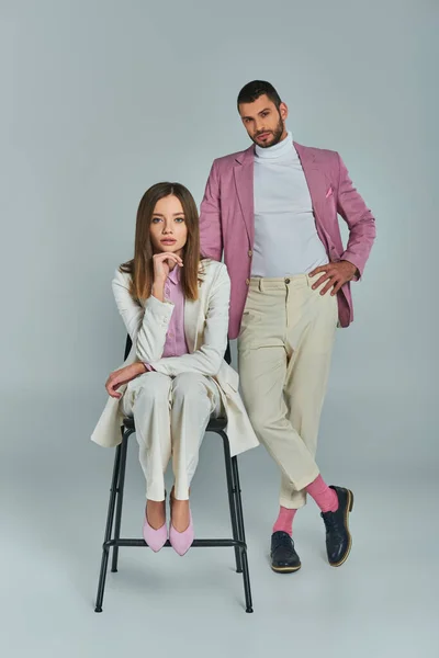 Hombre seguro en blazer lila con la mano en la cadera cerca de la mujer en traje blanco sentado en la silla en gris - foto de stock