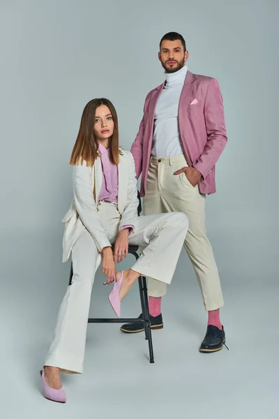 Stylish woman in white suit sitting on chair near confident man in lilac blazer on grey, full length — Stock Photo