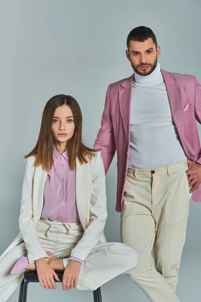 Modern and stylish business couple, man with hand on pocket near woman sitting on chair on grey — Stock Photo