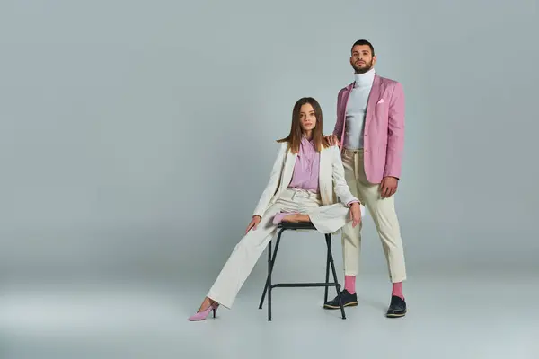 Elegant woman in white suit posing on chair near trendy man in lilac blazer on grey, full length — Stock Photo