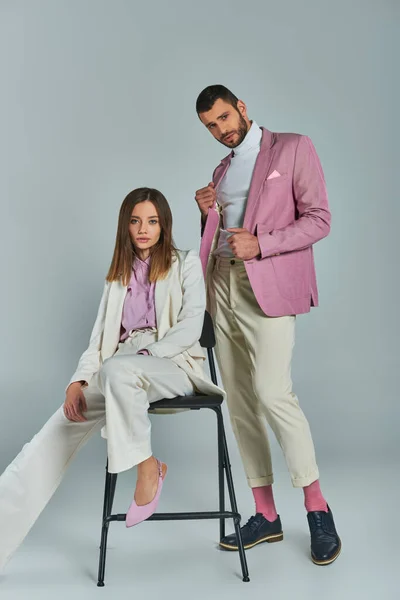 Trendy man in lilac blazer looking at camera near young woman in white suit sitting on chair on grey — Stock Photo