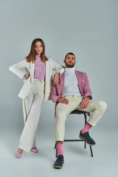 Woman in white formal suit posing with hand on hip near man in lilac blazer sitting on chair on grey — Stock Photo