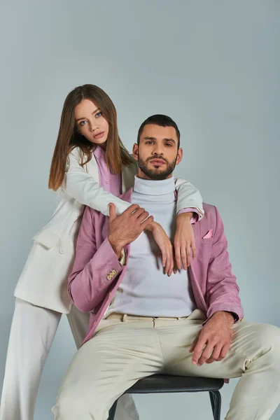 Trendy woman in white suit embracing confident man in lilac blazer sitting on chair on grey backdrop — Stock Photo