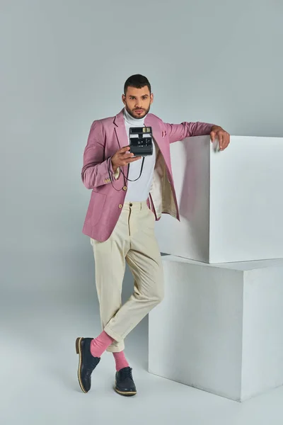 Stylish man in lilac blazer and white pants taking photo on vintage camera near cubes on grey — Stock Photo