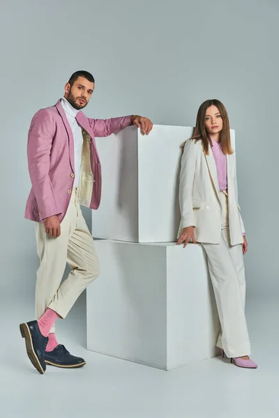 Молодая уверенная пара в модных костюмах позирует рядом с белыми кубиками на серой, минималистской моде — стоковое фото